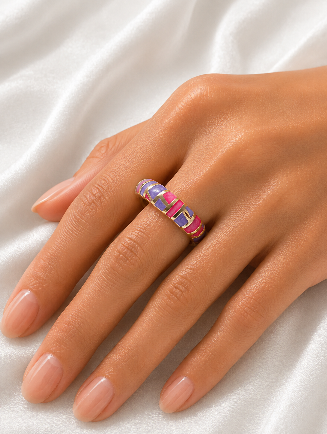 A close-up of a hand wearing a thin ring with a colorful striped pattern of pink, purple, and blue, resting on a white satin fabric background.