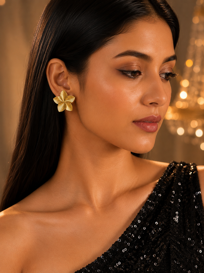 A woman wearing a single gold flower-shaped stud earring, shown in a close-up portrait with warm blurred lights in the background.
