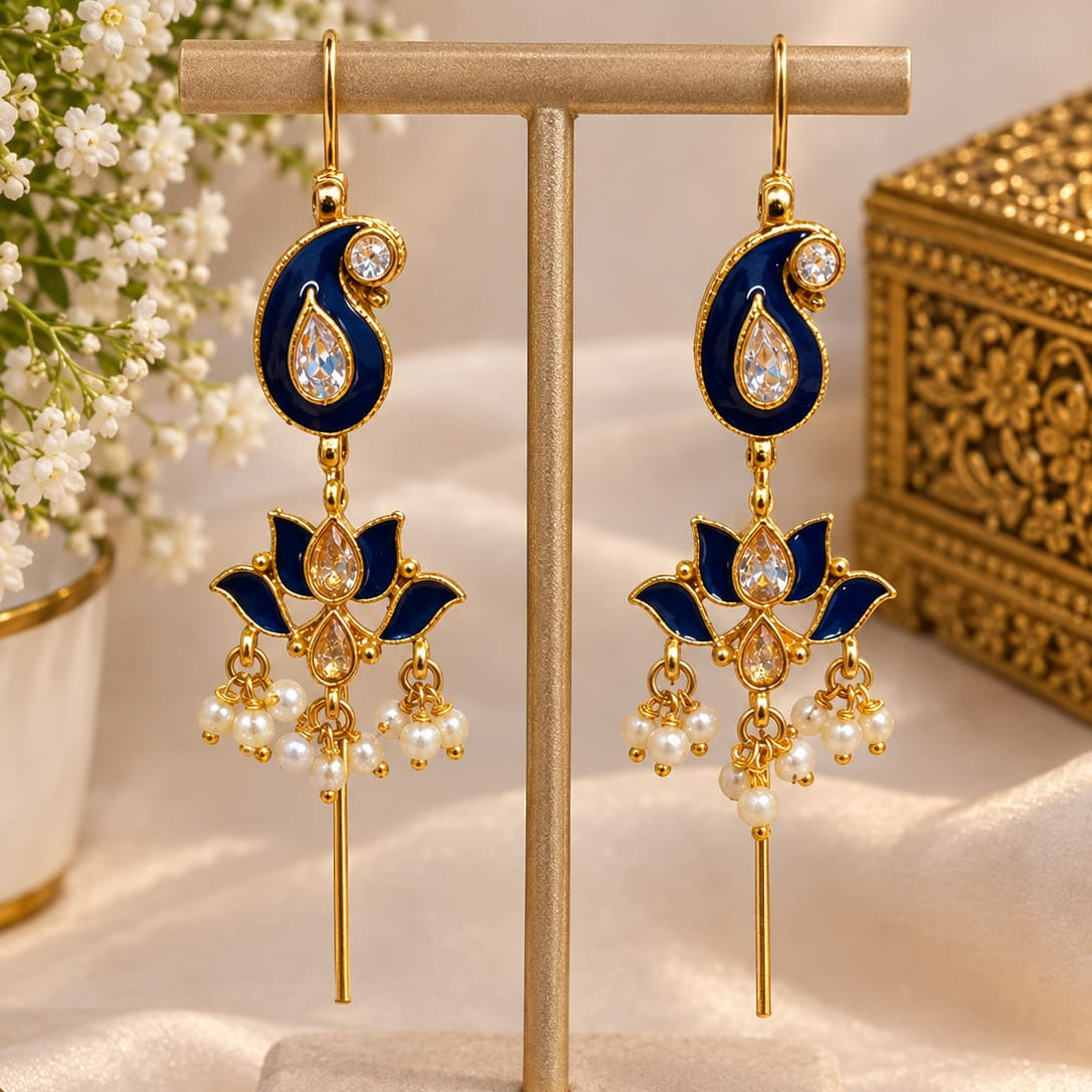 A pair of gold-plated Indian Kalka earrings with navy blue enamel, clear teardrop crystals, and white pearl bead drops, displayed on a beige jewelry stand.