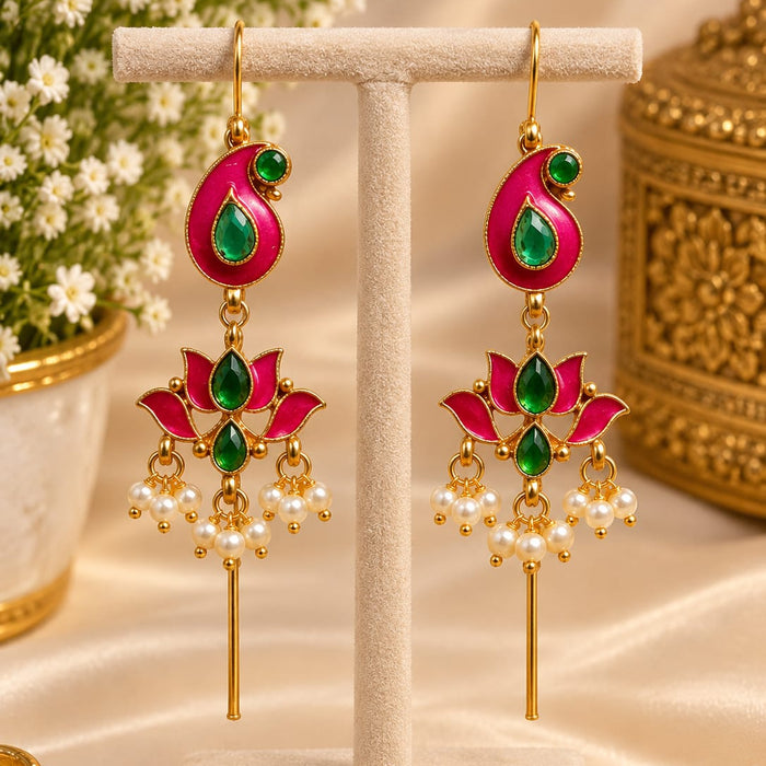 A pair of gold-toned Indian-style dangle earrings with bright pink and green enamel details, displayed on a cream-colored jewelry stand. The background features a soft beige satin fabric, white baby's breath flowers, and a decorative gold container.