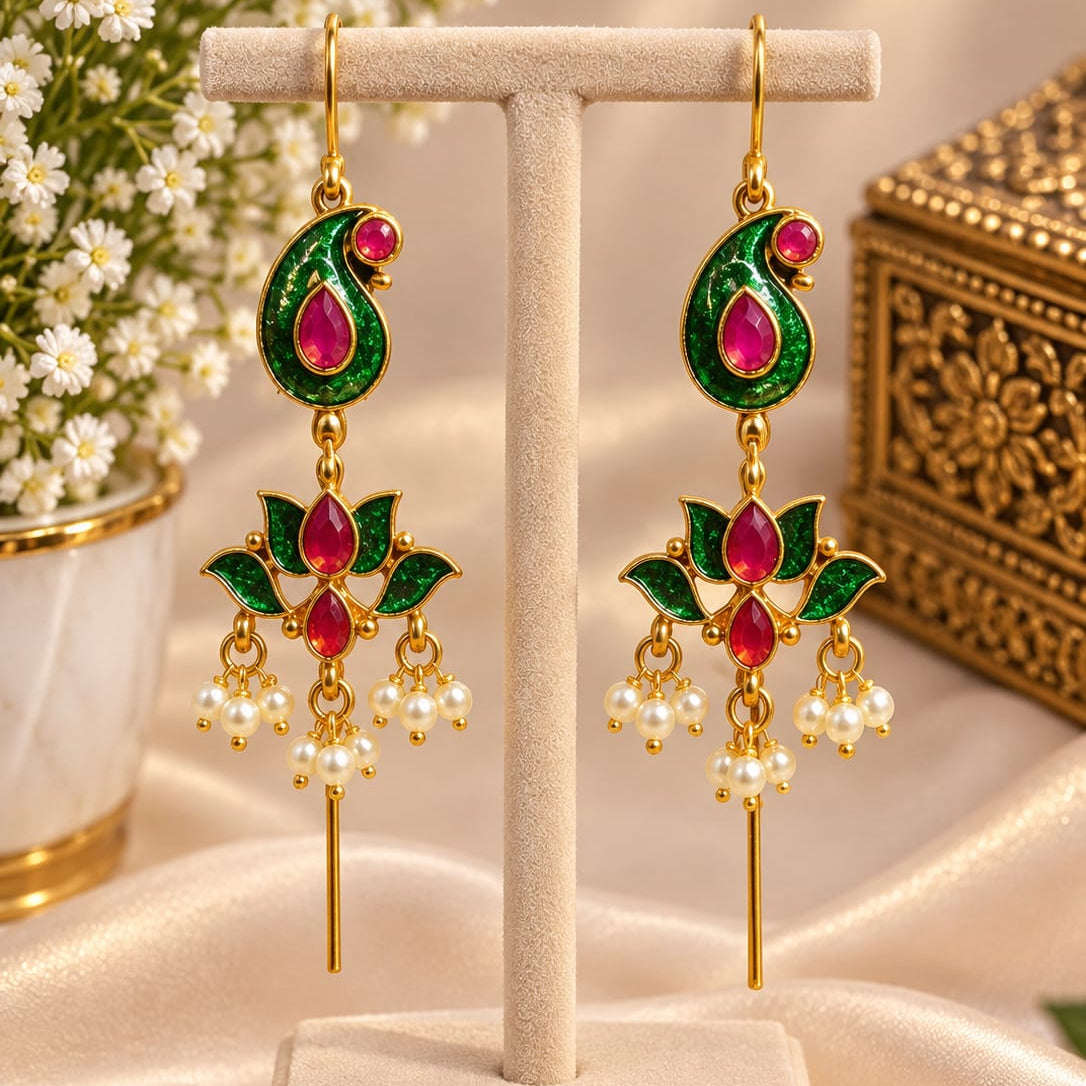 A pair of gold-plated Indian jhumka earrings with green enamel paisley tops and red stone accents, hanging on a beige jewelry display stand. The background features a soft beige satin fabric, white baby's breath flowers, and a decorative gold box.