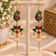 A pair of gold-plated Indian jhumka earrings with green enamel paisley tops and red stone accents, hanging on a beige jewelry display stand. The background features a soft beige satin fabric, white baby's breath flowers, and a decorative gold box.
