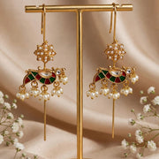 Gold-toned traditional Indian jhumka earrings with green and red enamel details, pearl bead drops, and a central stone, displayed on a gold stand against a beige fabric backdrop with baby's breath flowers.