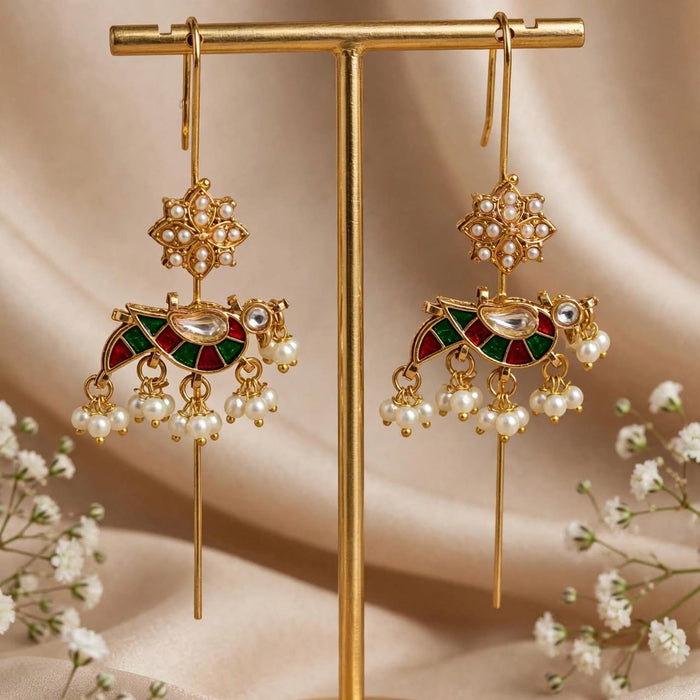 Gold-toned traditional Indian jhumka earrings with green and red enamel details, pearl bead drops, and a central stone, displayed on a gold stand against a beige fabric backdrop with baby's breath flowers.