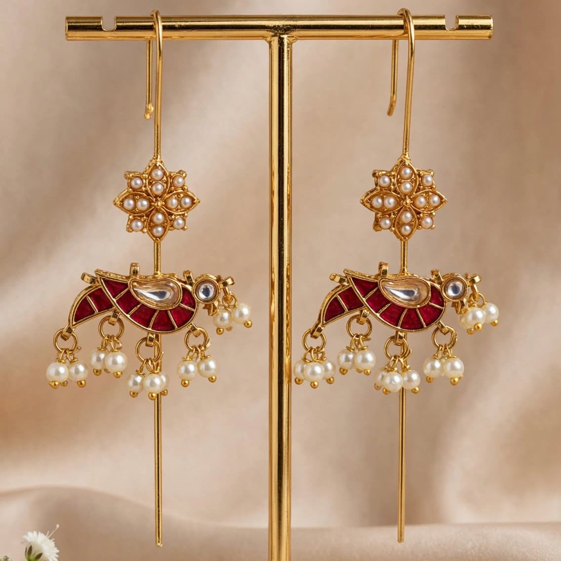 Gold-toned traditional Indian jhumka earrings with red enamel, white pearl bead drops, and small clear stone accents, displayed on a gold stand against a beige satin backdrop with small white flowers.