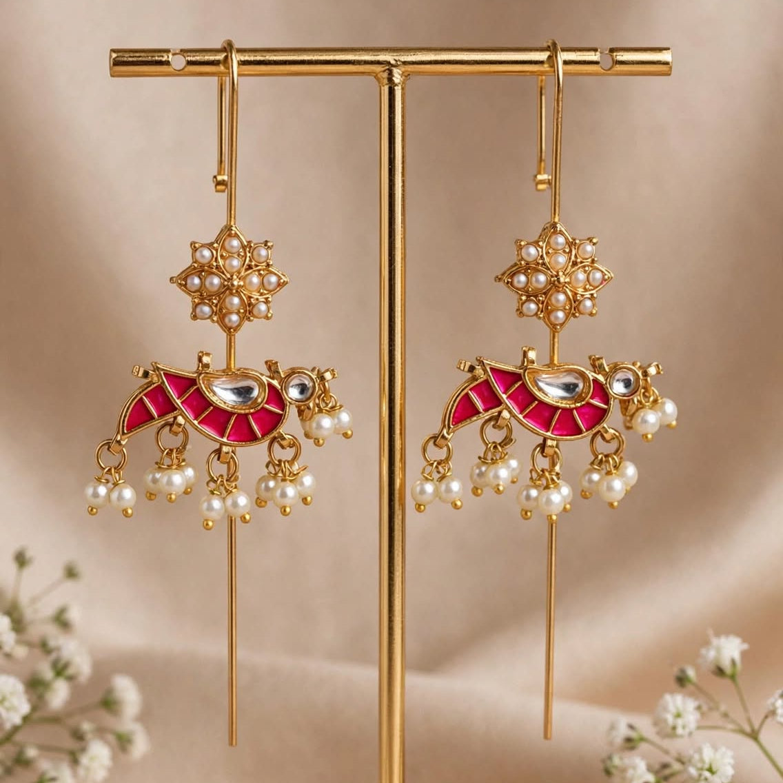 Gold-toned traditional Indian earrings with red enamel, pearl bead drops, and a floral motif, displayed on a gold stand with baby's breath flowers on a beige background.