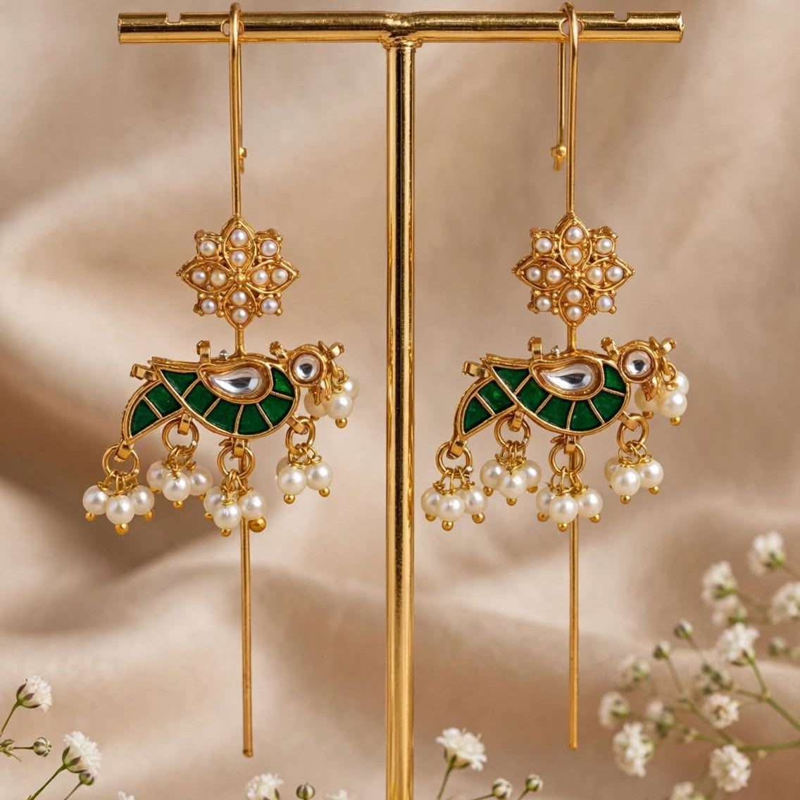 A pair of ornate gold-toned dangle earrings with green enamel, pearl-like beads, and dangling floral elements, displayed on a gold stand against a soft beige fabric backdrop with small white flowers around it.