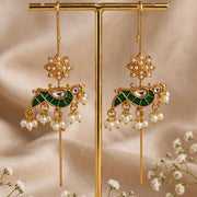 A pair of ornate gold-toned dangle earrings with green enamel, pearl-like beads, and dangling floral elements, displayed on a gold stand against a soft beige fabric backdrop with small white flowers around it.