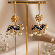 A pair of gold-toned dangle earrings with dark blue enamel details, floral motifs, and dangling pearl beads, displayed on a gold stand against a soft beige fabric backdrop with small white flowers.