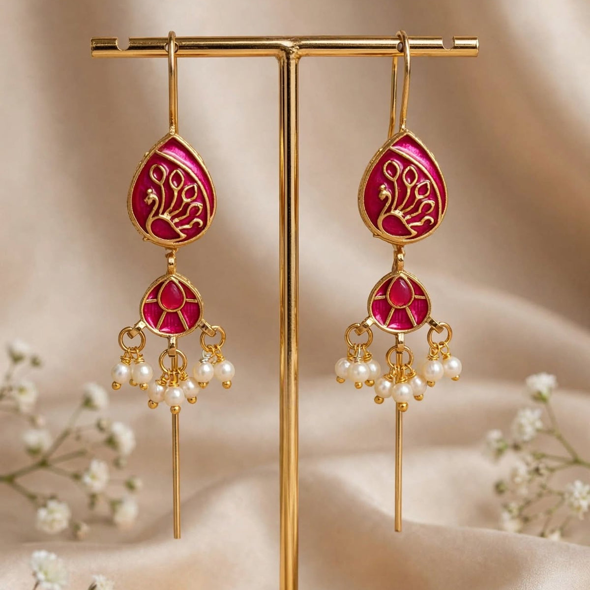Gold-toned dangle earrings with bright pink enamel panels and white pearl-like drops, displayed on a gold stand against a soft beige fabric backdrop with small white flowers.
