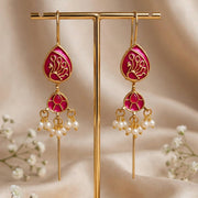 Gold-toned dangle earrings with bright pink enamel panels and white pearl-like drops, displayed on a gold stand against a soft beige fabric backdrop with small white flowers.