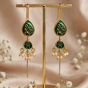 A pair of gold and green traditional Indian earrings displayed on a gold stand, surrounded by small white flowers on a beige fabric background.