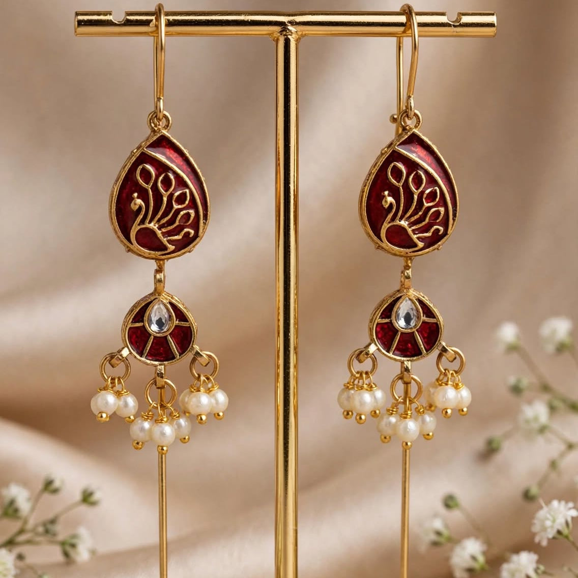 A pair of gold-toned traditional Indian dangle earrings with deep red enamel panels, gold floral engravings, and hanging pearl clusters, displayed on a gold stand against a beige satin background with small white flowers.