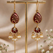 A pair of gold-toned traditional Indian dangle earrings with deep red enamel panels, gold floral engravings, and hanging pearl clusters, displayed on a gold stand against a beige satin background with small white flowers.