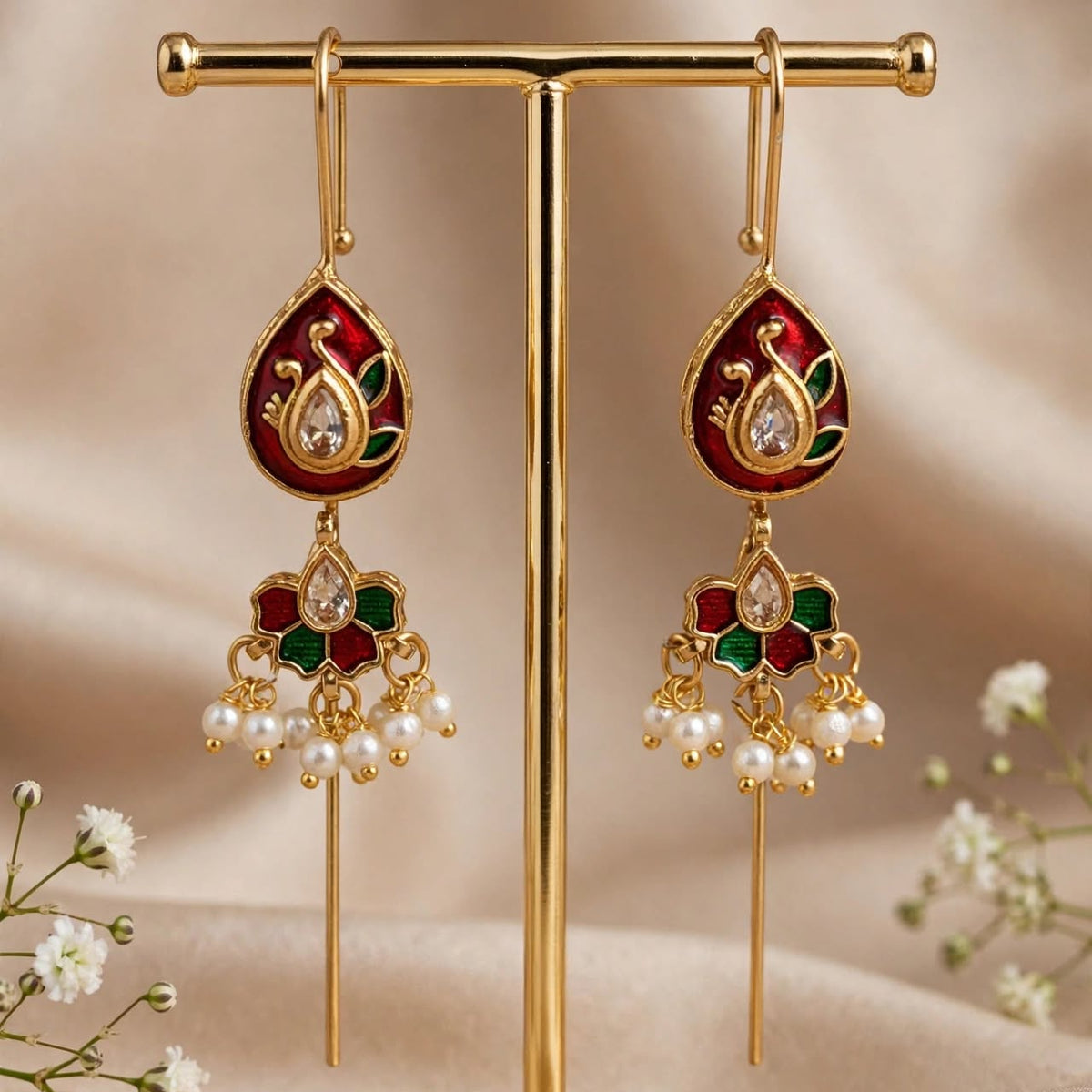 A pair of gold-toned Indian jhumka earrings with red, green, and white enamel details and pearl bead drops, displayed on a gold stand against a beige fabric backdrop.