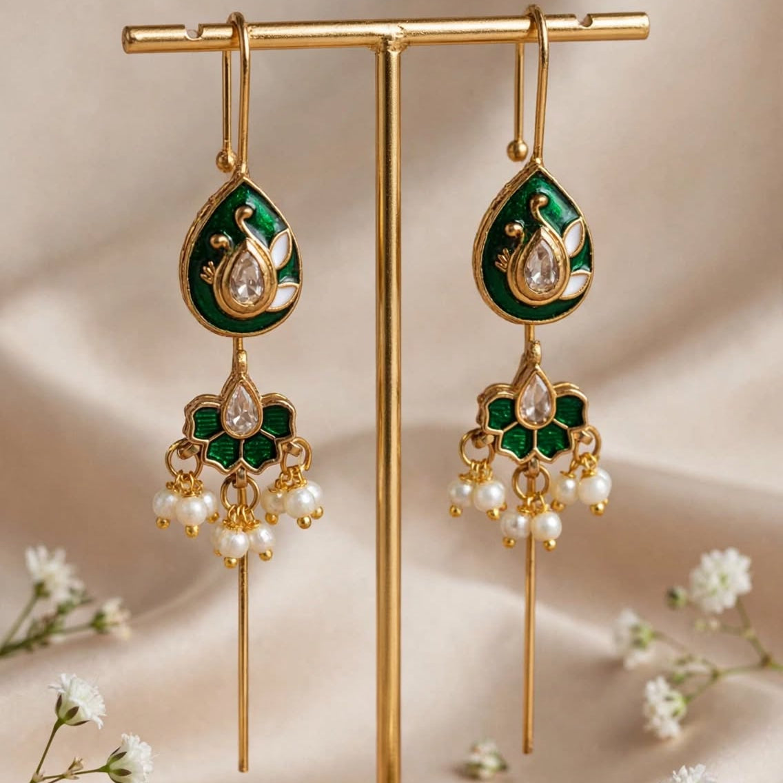 A pair of gold-tone and emerald green teardrop dangle earrings with white pearl-like drops, displayed on a gold stand against a beige satin backdrop with small white flowers.
