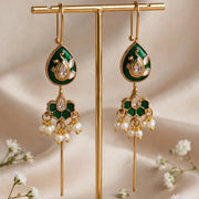 A pair of gold-tone and emerald green teardrop dangle earrings with white pearl-like drops, displayed on a gold stand against a beige satin backdrop with small white flowers.
