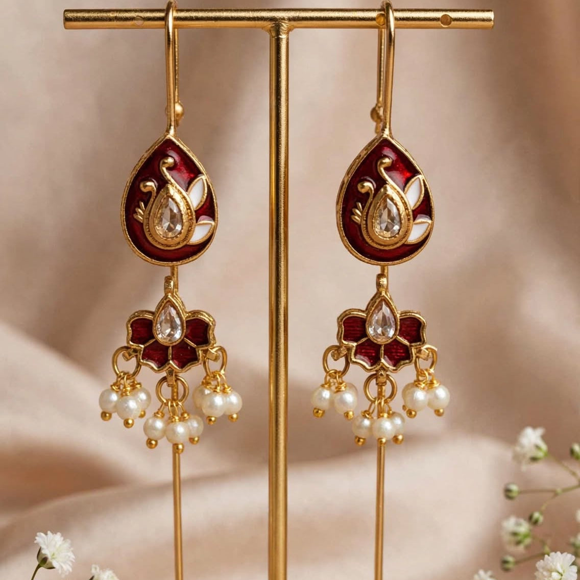 A pair of gold-toned traditional Indian earrings with deep red enamel and white pearl-like drops, displayed on a gold stand against a soft beige fabric backdrop with small white flowers.