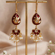 A pair of gold-toned traditional Indian earrings with deep red enamel and white pearl-like drops, displayed on a gold stand against a soft beige fabric backdrop with small white flowers.