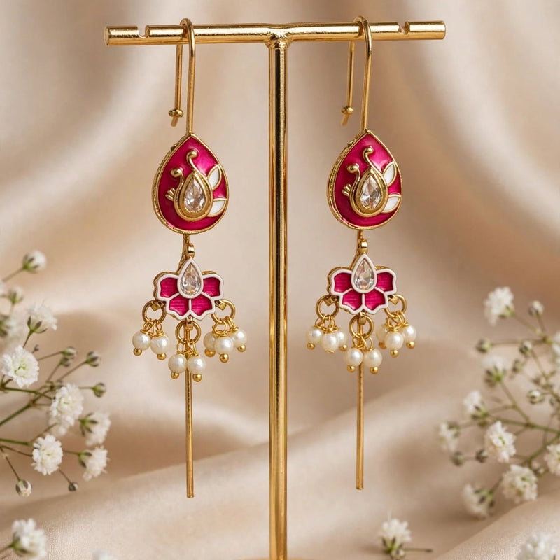 Gold-toned dangle earrings with bright pink enamel teardrop pendants and pearl-like drops, displayed on a gold stand against a soft beige satin backdrop with small white flowers.