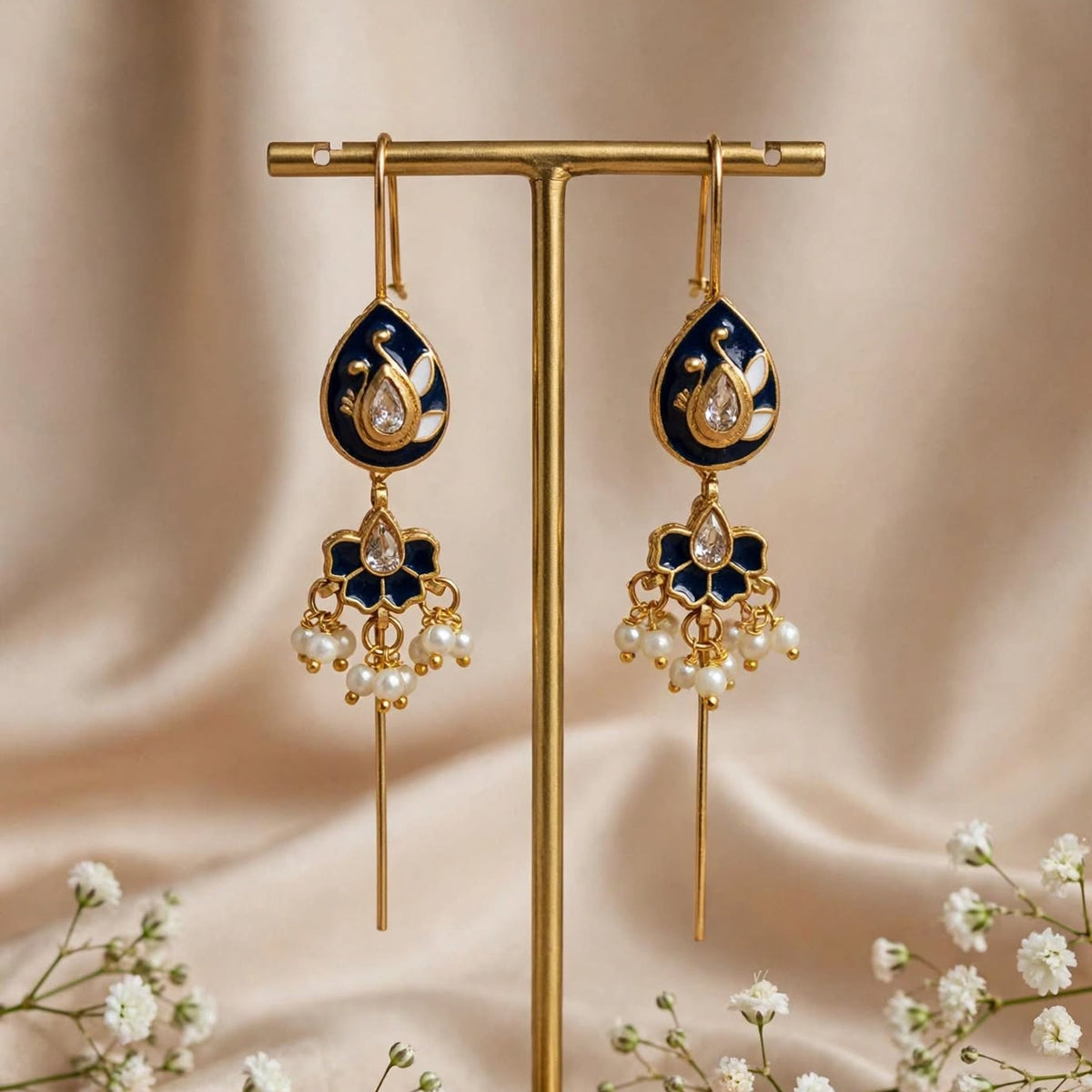 A pair of ornate gold-tone dangle earrings with dark blue enamel and clear teardrop stones, displayed on a gold stand against a soft beige satin backdrop with small white flowers.