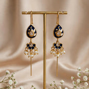 A pair of ornate gold-tone dangle earrings with dark blue enamel and clear teardrop stones, displayed on a gold stand against a soft beige satin backdrop with small white flowers.