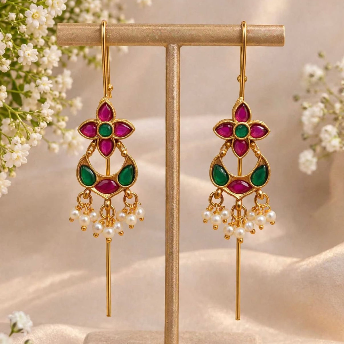 A pair of gold-tone dangle earrings with magenta and green stone accents and pearl-like drops, displayed on a beige jewelry stand with soft floral props and draped fabric in the background.