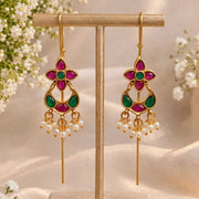 A pair of gold-tone dangle earrings with magenta and green stone accents and pearl-like drops, displayed on a beige jewelry stand with soft floral props and draped fabric in the background.