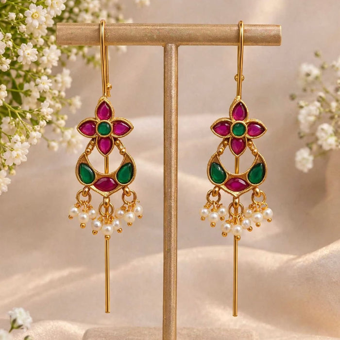 A pair of gold-tone dangle earrings with magenta and green stone accents and pearl-like drops, displayed on a beige jewelry stand with soft floral props and draped fabric in the background.