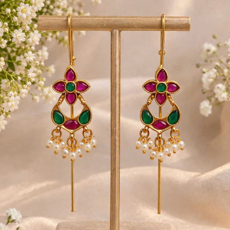 A pair of gold-tone dangle earrings with magenta and green stone accents and pearl-like drops, displayed on a beige jewelry stand with soft floral props and draped fabric in the background.