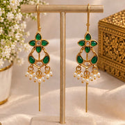 A pair of gold-toned traditional Indian chandelier earrings with green stone accents and white pearl drops, displayed on a beige jewelry stand.