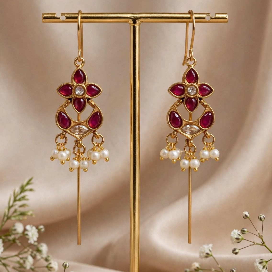 Gold-toned traditional dangle earrings with deep red stones and small pearl-like drops, displayed on a gold stand against a soft beige satin backdrop with white flowers at the base.