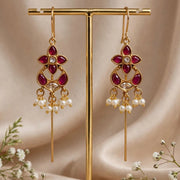 Gold-toned traditional dangle earrings with deep red stones and small pearl-like drops, displayed on a gold stand against a soft beige satin backdrop with white flowers at the base.